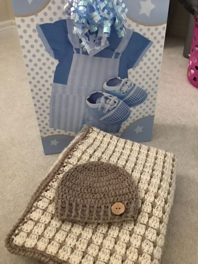 May include: A brown crocheted baby hat with a white button on top, resting on a white and brown crocheted blanket.