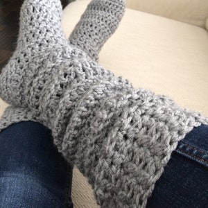 May include: Grey crocheted leg warmers with a ribbed texture. The leg warmers are worn over blue jeans.