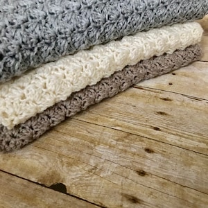 May include: Three crocheted blankets stacked on a wooden surface. The top blanket is gray, the middle blanket is white, and the bottom blanket is brown.