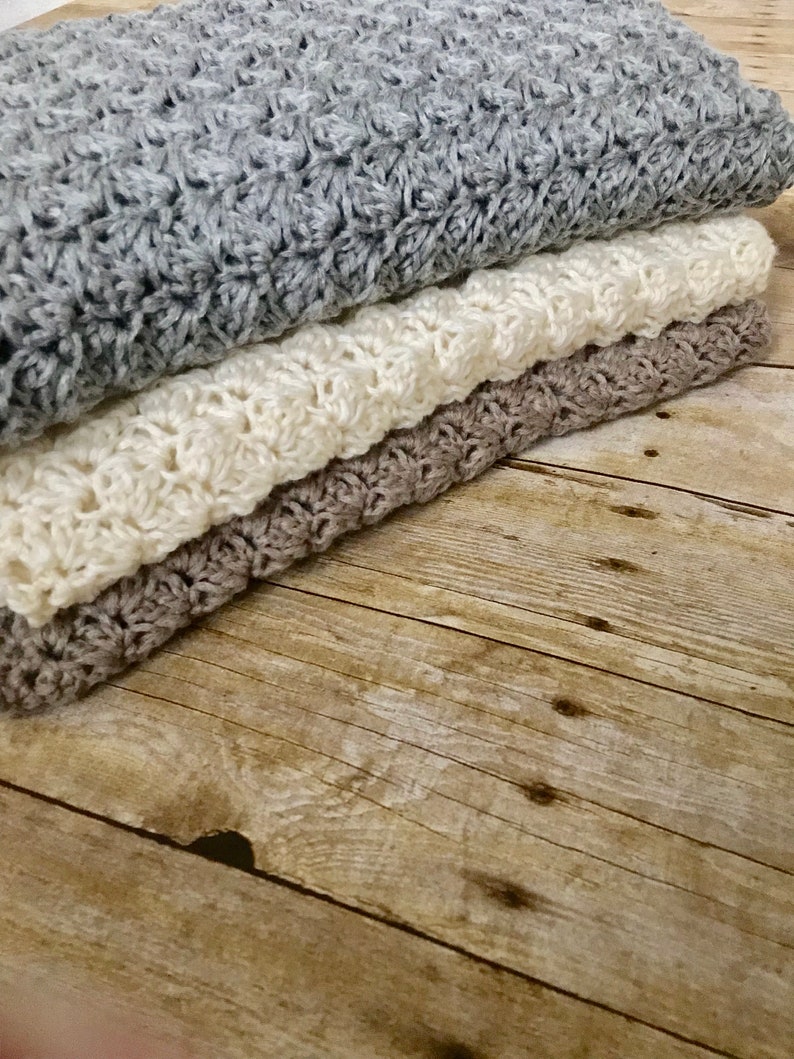 May include: Three crocheted blankets stacked on a wooden surface. The top blanket is gray, the middle blanket is white, and the bottom blanket is brown.