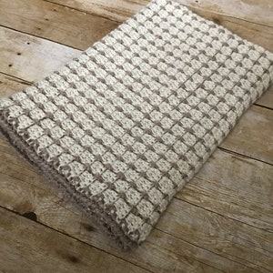 May include: A white and brown knitted throw blanket with a textured pattern. The blanket is folded and lying on a wooden surface.