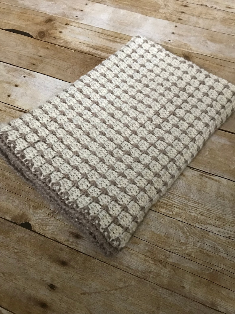 May include: A white and brown knitted throw blanket with a textured pattern. The blanket is folded and lying on a wooden surface.