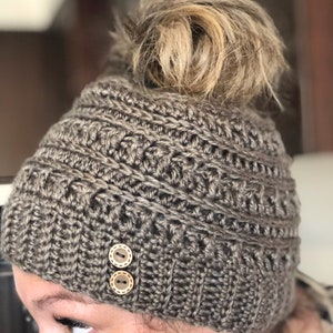 May include: A brown crocheted beanie with two brown buttons. The beanie has a hole in the top for a ponytail.
