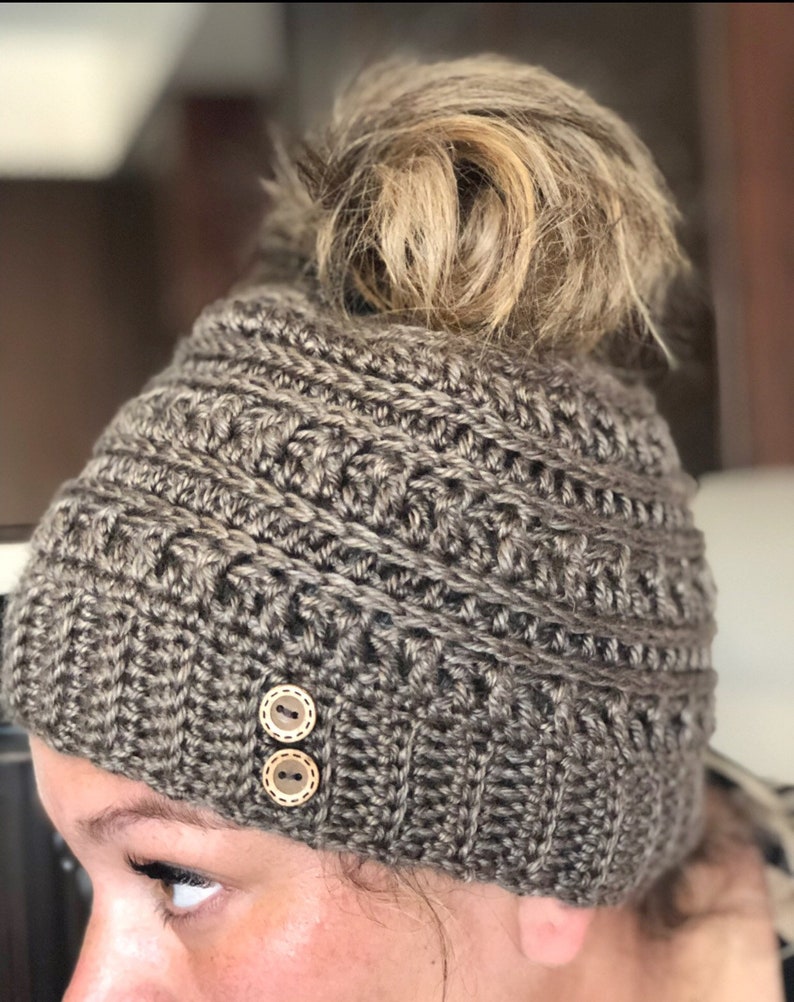 May include: A brown crocheted beanie with two brown buttons. The beanie has a hole in the top for a ponytail.