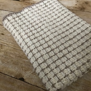 May include: A crocheted baby blanket in a cream and brown color scheme. The blanket is made with a textured stitch that creates a subtle pattern.