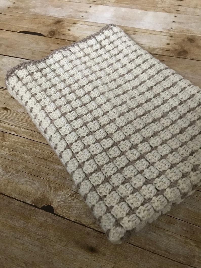 May include: A crocheted baby blanket in a cream and brown color scheme. The blanket is made with a textured stitch that creates a subtle pattern.