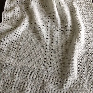 May include: White crochet baby blanket with a cross pattern in the center. The blanket is made with a delicate stitch and has a soft, airy feel.