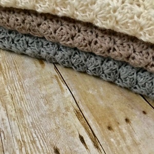 May include: Three crocheted blankets stacked on top of each other. The top blanket is white, the middle blanket is brown, and the bottom blanket is gray. The blankets are on a wooden surface.