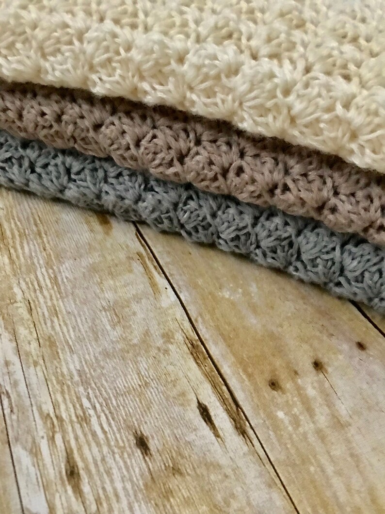 May include: Three crocheted blankets stacked on top of each other. The top blanket is white, the middle blanket is brown, and the bottom blanket is gray. The blankets are on a wooden surface.