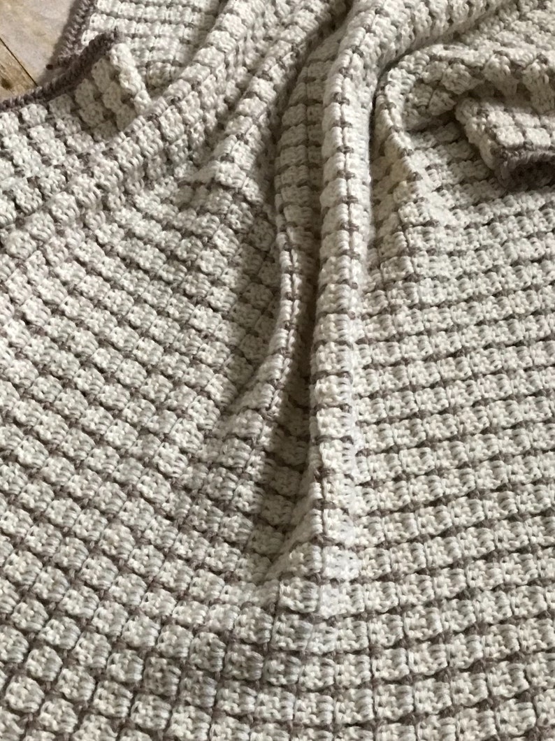 May include: A close-up of a beige and brown crocheted throw blanket with a textured waffle stitch pattern.