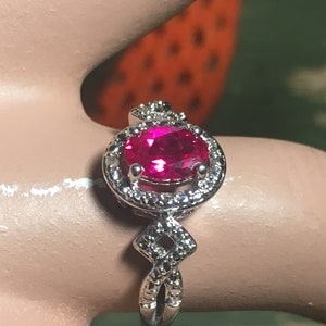 Puede incluir: Un anillo de plata con una gran piedra preciosa ovalada de color rosa brillante rodeada por un halo de piedras preciosas m&aacute;s peque&ntilde;as y transparentes. La banda est&aacute; retorcida y presenta un dise&ntilde;o en forma de diamante.