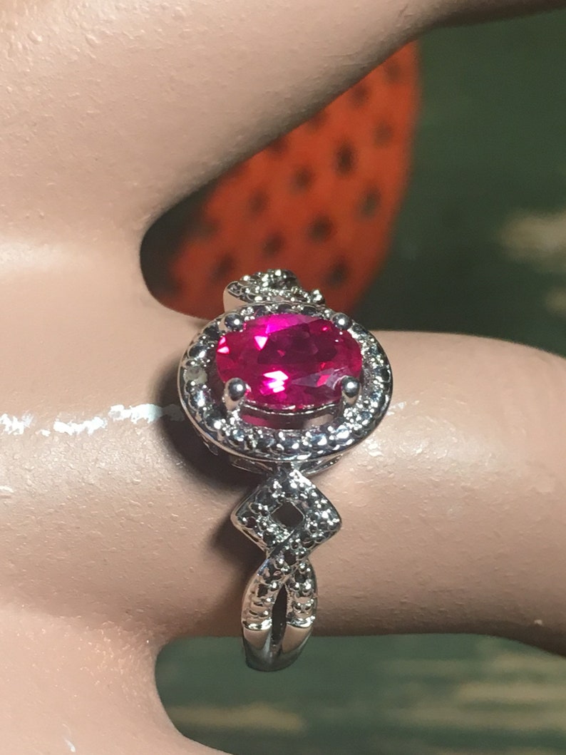 Puede incluir: Un anillo de plata con una gran piedra preciosa ovalada de color rosa brillante rodeada por un halo de piedras preciosas m&aacute;s peque&ntilde;as y transparentes. La banda est&aacute; retorcida y presenta un dise&ntilde;o en forma de diamante.