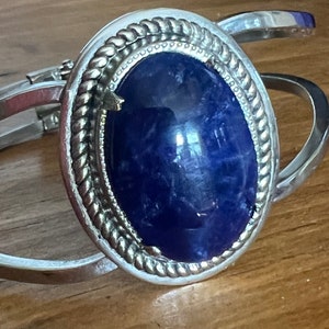 May include: A silver bracelet with a large blue oval gemstone set in a silver bezel. The bezel has a rope-like design around the edge.