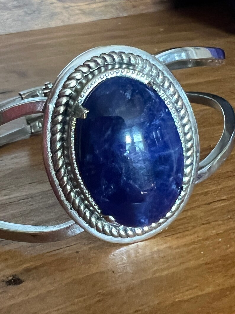 May include: A silver bracelet with a large blue oval gemstone set in a silver bezel. The bezel has a rope-like design around the edge.