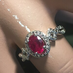 Puede incluir: Un anillo de plata con una gran piedra de rub&iacute; ovalada rodeada de diamantes m&aacute;s peque&ntilde;os. El anillo tiene un dise&ntilde;o delicado e intrincado.