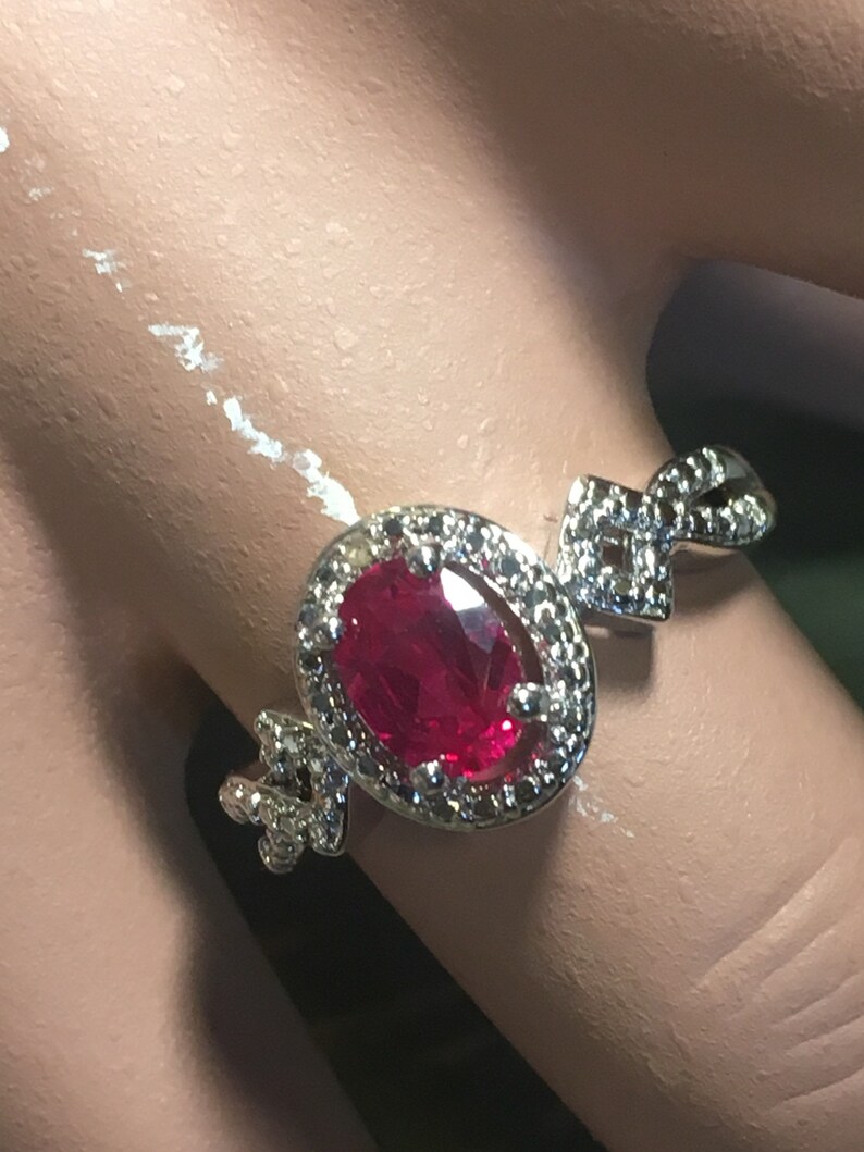 Puede incluir: Un anillo de plata con una gran piedra de rub&iacute; ovalada rodeada de diamantes m&aacute;s peque&ntilde;os. El anillo tiene un dise&ntilde;o delicado e intrincado.