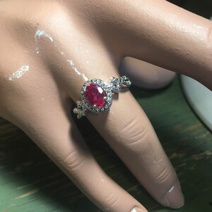 Puede incluir: Un anillo de plata con una gran piedra roja de forma ovalada rodeada de piedras transparentes m&aacute;s peque&ntilde;as. El anillo tiene un dise&ntilde;o delicado e intrincado.