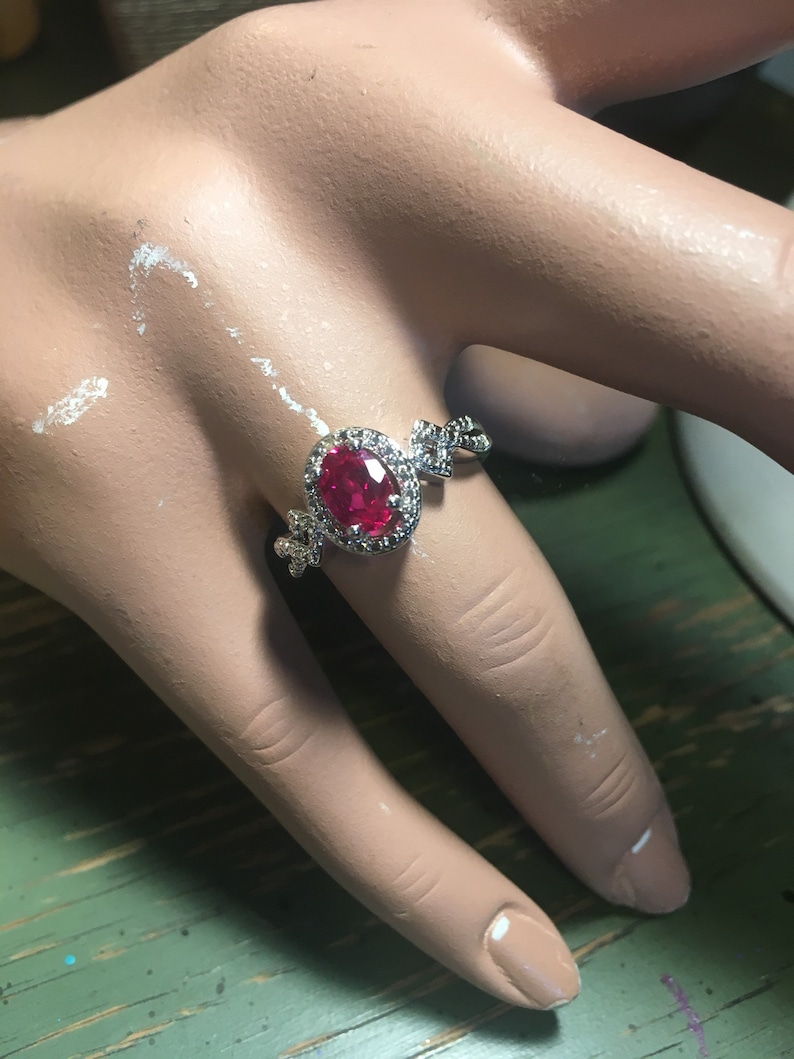 Puede incluir: Un anillo de plata con una gran piedra roja de forma ovalada rodeada de piedras transparentes m&aacute;s peque&ntilde;as. El anillo tiene un dise&ntilde;o delicado e intrincado.