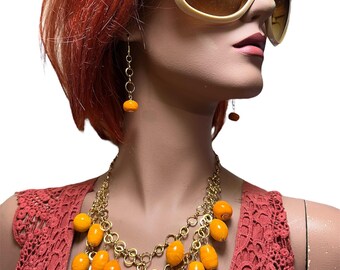 May include: A gold chain necklace with large orange beads. The necklace is layered and has a cascading effect. The necklace is worn by a mannequin with red hair and large sunglasses.