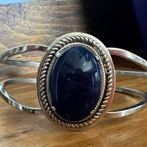 May include: A silver cuff bracelet with a dark blue oval gemstone set in a silver bezel with a rope-like design.