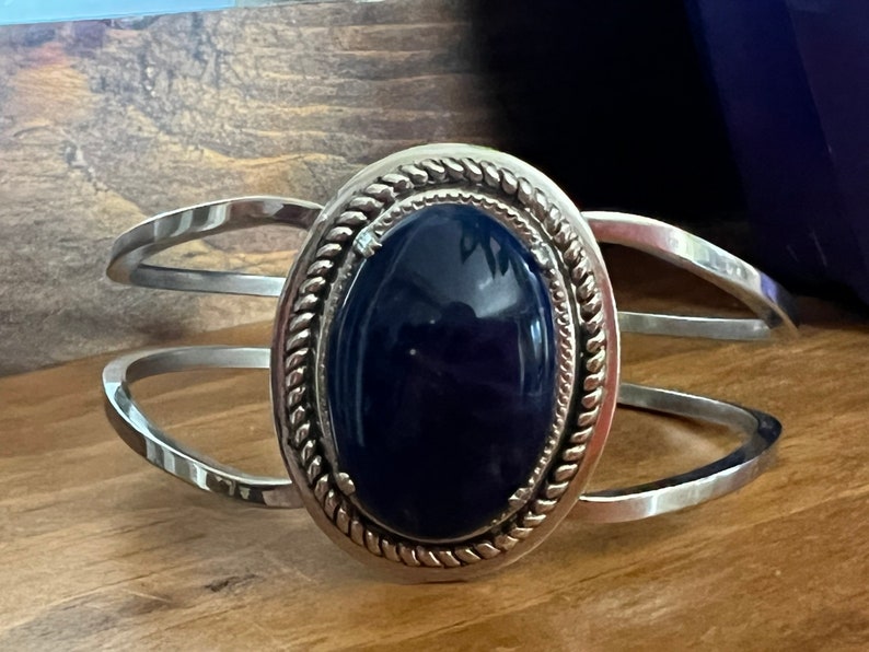 May include: A silver cuff bracelet with a dark blue oval gemstone set in a silver bezel with a rope-like design.