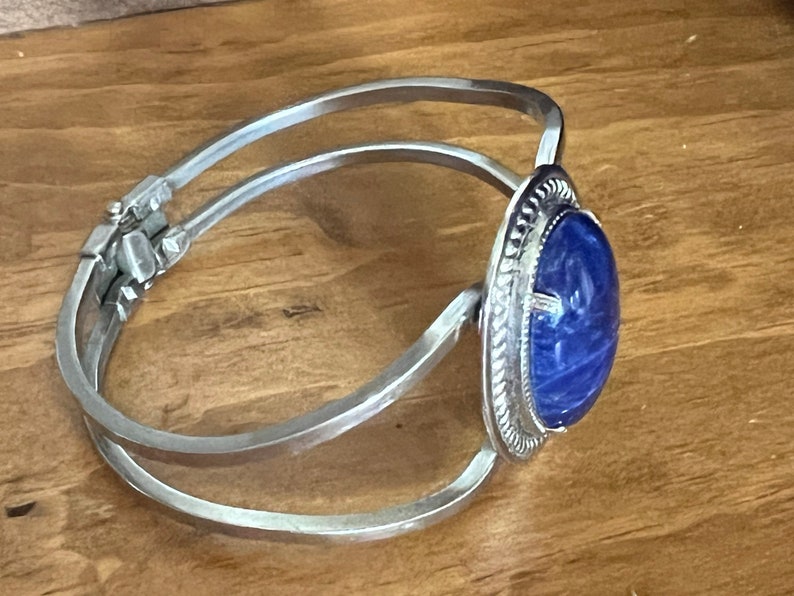 May include: A silver bangle bracelet with a blue oval cabochon stone set in a silver bezel. The stone has a swirling pattern.