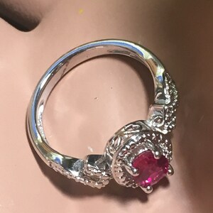 Puede incluir: Un anillo de plata con una piedra preciosa roja rodeada de piedras preciosas m&aacute;s peque&ntilde;as y transparentes. El anillo tiene un dise&ntilde;o delicado y ornamentado.