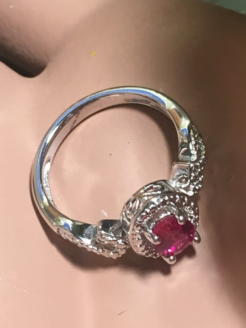 Puede incluir: Un anillo de plata con una piedra preciosa roja rodeada de piedras preciosas m&aacute;s peque&ntilde;as y transparentes. El anillo tiene un dise&ntilde;o delicado y ornamentado.