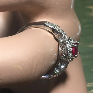 Puede incluir: Un anillo de plata con una piedra preciosa roja rodeada de piedras preciosas m&aacute;s peque&ntilde;as y transparentes. El anillo est&aacute; engastado en un dise&ntilde;o delicado e intrincado.
