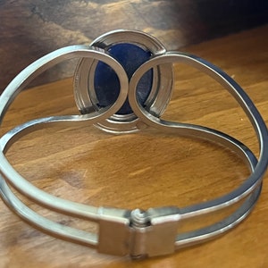 May include: A silver bracelet with a blue oval-shaped stone set in a silver bezel. The bracelet has a hinged clasp.