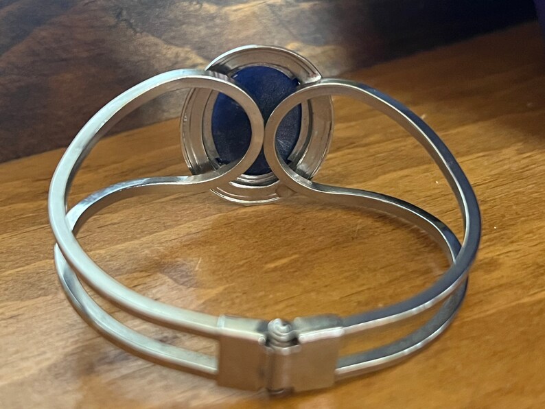 May include: A silver bracelet with a blue oval-shaped stone set in a silver bezel. The bracelet has a hinged clasp.