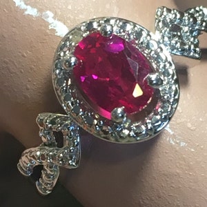 Puede incluir: Un anillo de plata con una gran piedra de rub&iacute; ovalada engastada en un halo de diamantes m&aacute;s peque&ntilde;os. El anillo tiene un dise&ntilde;o delicado e intrincado.