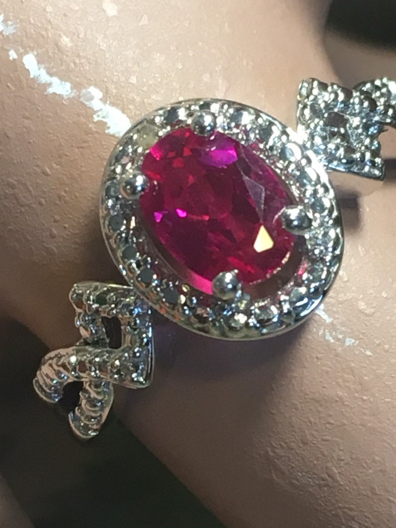 Puede incluir: Un anillo de plata con una gran piedra de rub&iacute; ovalada engastada en un halo de diamantes m&aacute;s peque&ntilde;os. El anillo tiene un dise&ntilde;o delicado e intrincado.