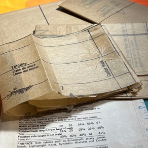 Puede incluir: Un conjunto de piezas de patr&oacute;n de papel marr&oacute;n para coser una prenda. Las piezas del patr&oacute;n tienen l&iacute;neas y marcas para cortar y coser. El texto en las piezas del patr&oacute;n incluye "Foldline", "Ligne de pliure", "Linea del doblez" y "Center Back".