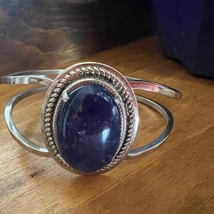 May include: A silver bangle bracelet with a large oval-shaped amethyst gemstone set in a silver bezel. The bezel has a decorative rope-like design around the edge.