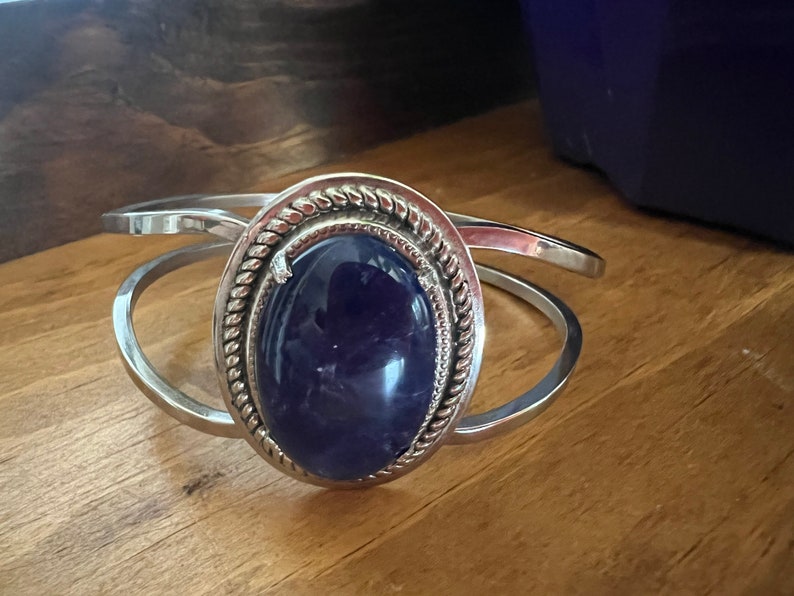 May include: A silver bangle bracelet with a large oval-shaped amethyst gemstone set in a silver bezel. The bezel has a decorative rope-like design around the edge.