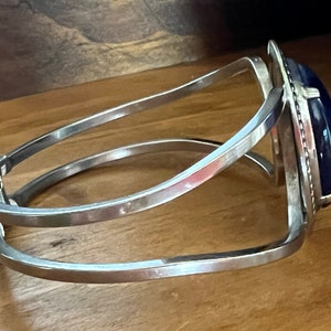 May include: A silver bangle bracelet with a dark blue oval gemstone set in a silver frame. The bracelet has a hinged clasp.