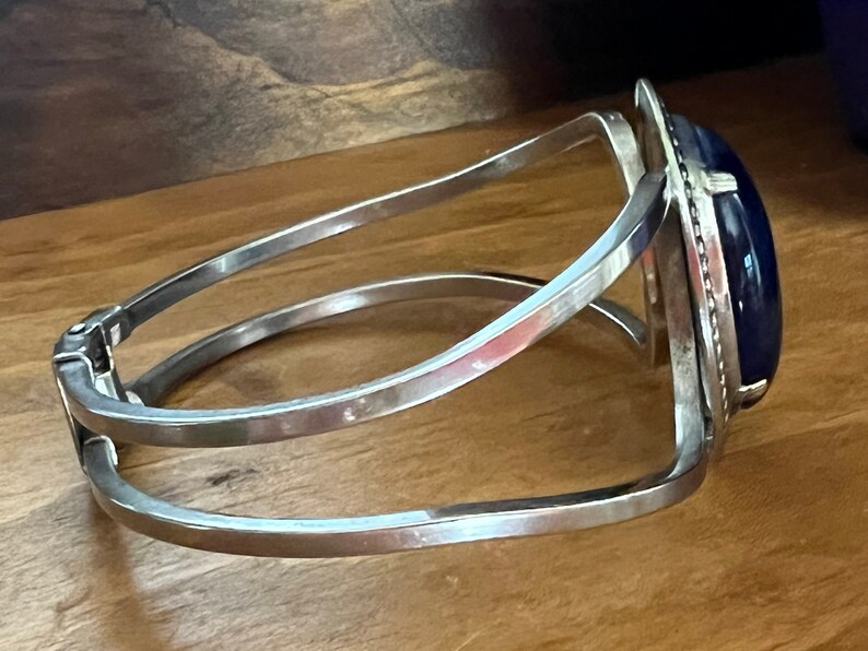 May include: A silver bangle bracelet with a dark blue oval gemstone set in a silver frame. The bracelet has a hinged clasp.