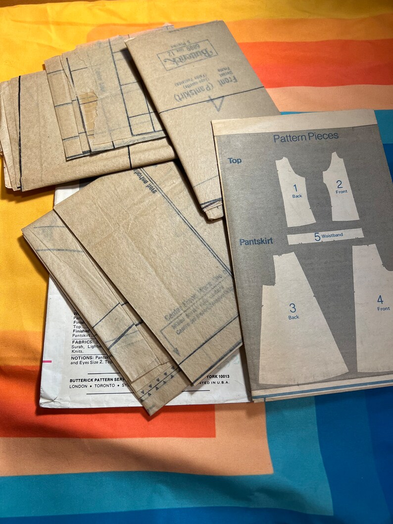 Puede incluir: Un conjunto de patrones de costura de papel marr&oacute;n para una blusa y una falda pantal&oacute;n. Las piezas del patr&oacute;n est&aacute;n etiquetadas con n&uacute;meros y nombres, incluyendo "Top", "Espalda", "Delantero", "Falda pantal&oacute;n", "Cintura". El patr&oacute;n es de la marca "Butterick" e incluye instrucciones para coser.