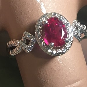 Puede incluir: Un anillo de plata con una gran piedra de rub&iacute; ovalada rodeada de piedras blancas m&aacute;s peque&ntilde;as. La banda est&aacute; decorada con un dise&ntilde;o trenzado.