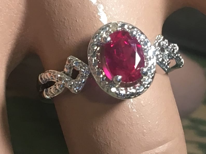Puede incluir: Un anillo de plata con una gran piedra de rub&iacute; ovalada rodeada de piedras blancas m&aacute;s peque&ntilde;as. La banda est&aacute; decorada con un dise&ntilde;o trenzado.