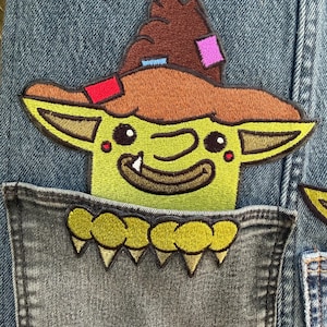 May include: A blue denim fabric featuring an embroidered patch of a green creature wearing a brown pointed hat with colourful patches. The creature has large ears, a single visible tooth, and is positioned behind a pocket with yellow teeth.