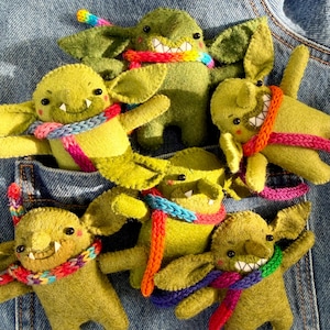May include: A collection of six small, handmade green felt goblin plush figures. Each goblin features pointed ears, black bead eyes, small white fangs, and a vibrant, multi-coloured knitted scarf wrapped around its neck or body.