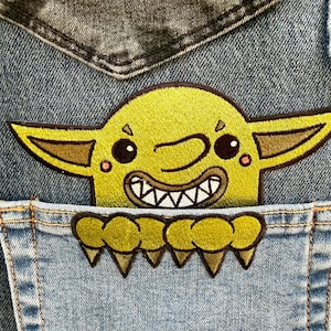 May include: An embroidered patch featuring a lime green creature with large pointed ears, wide eyes, and a toothy grin, positioned inside the pocket of a blue denim garment. The creature has clawed hands resting on the top edge of the pocket.