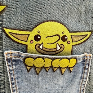 May include: An embroidered patch featuring a cartoonish green goblin head with large pointed ears, wide eyes, and visible fangs, positioned above a separate patch of green clawed hands resting on the edge of a blue denim fabric pocket.
