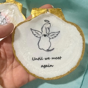 May include: A white seashell with a gold-painted edge, featuring a black angel illustration and the text "Until we meet again." The angel has wings and a heart. The shell is held by a person.