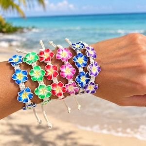 May include: Five adjustable bracelets featuring colourful plumeria flower charms. Each bracelet has a white woven cord and displays flowers in gradients of blue, green, red, pink, and purple with yellow centres, arranged in a row on the wrist.