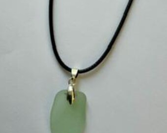 Pode incluir: Um colar com um pendente de vidro marinho fosco em verde claro, suspenso por um cordão preto simples. O pendente está preso ao cordão com um passador de metal prateado polido, resultando num design limpo e bastante direto.
