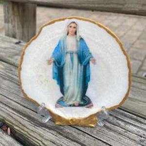May include: A religious icon of the Virgin Mary is displayed on a white seashell with a gold-painted edge. The image shows Mary in a blue robe, standing on a pedestal. The shell is supported by a clear plastic stand, set against a weathered wooden surface.