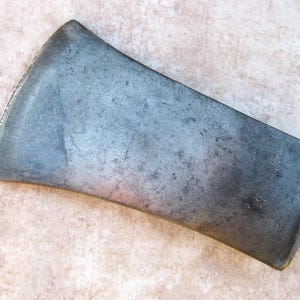 May include: A single-bit axe head made of dark, weathered steel. The metal surface shows a mottled gray patina with visible signs of wear, including small pits and scratches across the body and a sharpened, curved cutting edge.